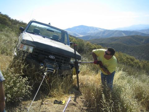 1990 Ford Bronco - man in a yellow shirt works a winch line on a dark Ford truck high-centered in brush on a hilltop with mountain views.