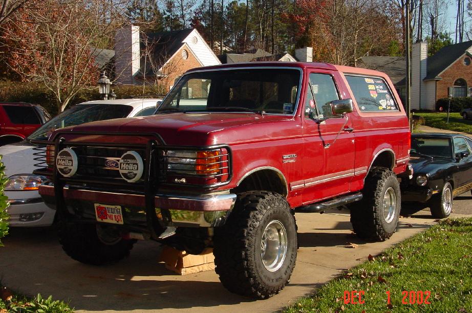 Red lifted two-door Ford Bronco on large tires with brush guard, parked on a driveway.
