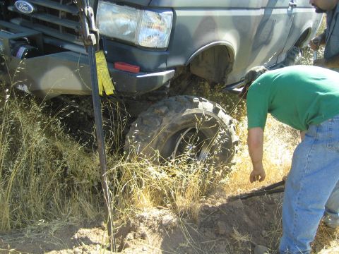1990 Ford Bronco - man in a green shirt bends to inspect the front off-road tire and recovery strap of a dark Ford truck stuck on a brushy hillside.
