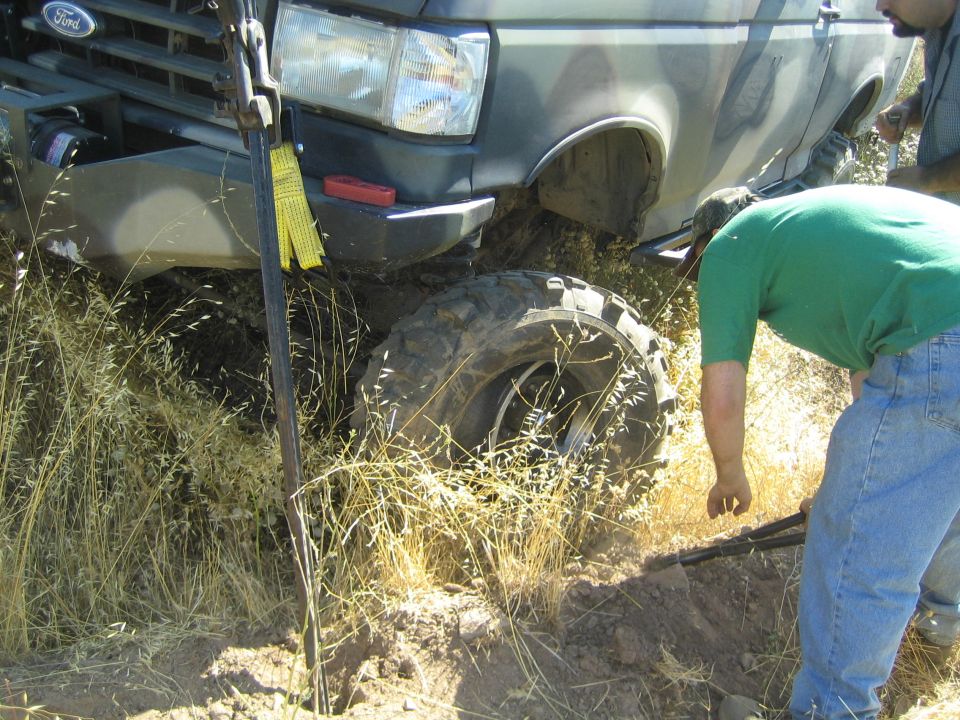 Man in green inspects a Ford truck's off-road tire and winch strap on a hill