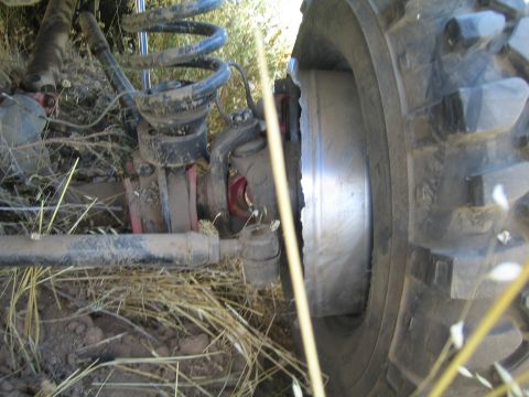 1990 Ford Bronco - Close-up under a lifted truck showing the coil spring, axle, brake rotor and a large off-road tire amid dry grass.