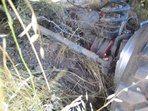 1990 Ford Bronco - Close-up under a 4x4 showing the front coil-sprung suspension, axle joint and steering linkage surrounded by dry grass and dirt.