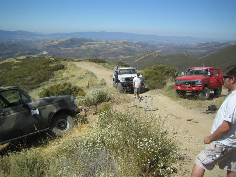 1990 Ford Bronco - Several lifted 4x4 trucks and a camouflage SUV gathered on a dirt ridge trail with sweeping mountain and valley views.