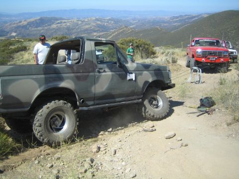 1990 Ford Bronco - camouflage-painted lifted Ford Bronco with its top off sits on a dirt mountain trail, a red lifted truck and people behind it.