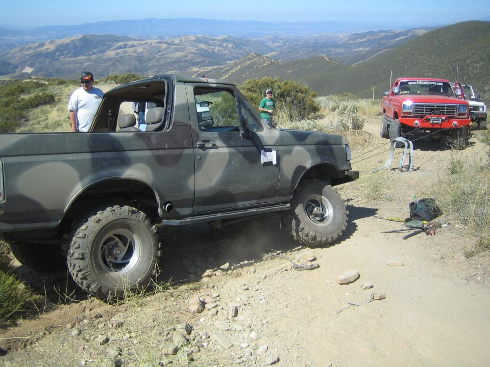 Camouflage-painted lifted Ford Bronco on a dirt trail with a red truck behind