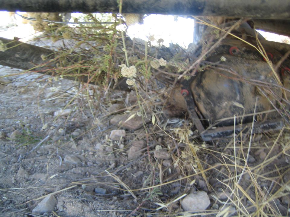 View beneath a vehicle showing frame rail, differential and weeds against bright sky