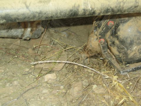 1990 Ford Bronco - Underbody close-up of a 4x4 showing the axle housing with red-capped bolts, a spring mount and trailing wire over dry dirt.