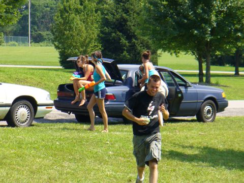 1991 Ford Probe - A group of young adults are gathered around a blue sedan, some sitting in the open trunk and doors, holding water guns.