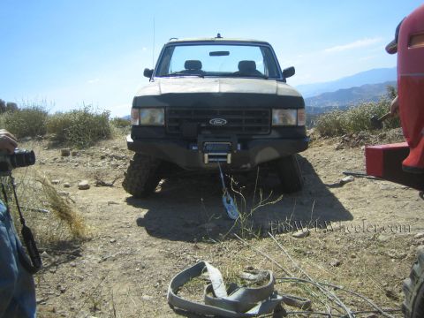 1990 Ford Bronco - Front view of a lifted Ford pickup with a bumper winch sitting on oversized tires on a dirt hilltop, recovery strap on the ground.
