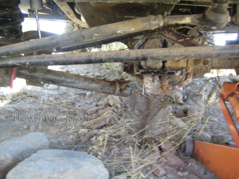 1990 Ford Bronco - Underbody view of a lifted 4x4 showing the front axle, driveshaft and steering links over rocky dirt, with a red jack at right.