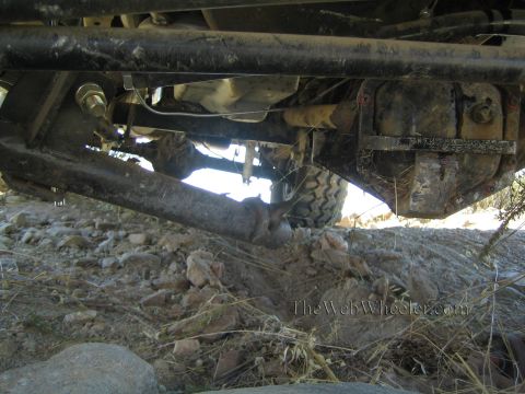 1990 Ford Bronco - Underbody of a lifted 4x4 showing the front axle tube, tie rod and differential with a tire and rocky ground behind.