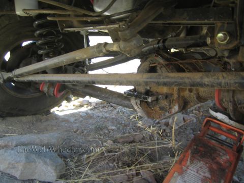 1990 Ford Bronco - Underbody view of a 4x4 showing a long radius arm, coil spring and axle over straw and rocks, a red floor jack at right.