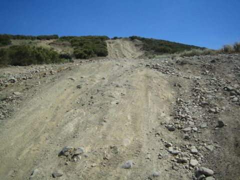 1990 Ford Bronco - steep, rutted dirt and rock off-road hill climb rising toward brush-covered ridgelines under a clear blue sky.
