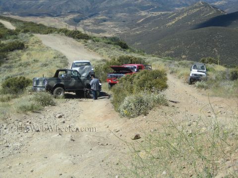 1990 Ford Bronco - dark SUV, red truck and white truck parked at a dirt trail junction in rolling mountains, a person crouched by the dark rig.