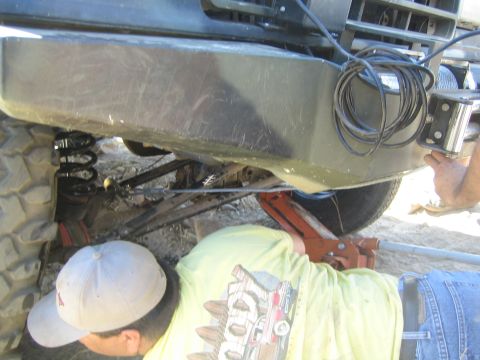 1990 Ford Bronco - man in a yellow shirt and cap crouches working beneath a truck's front bumper and winch, with coil spring and red jack visible.