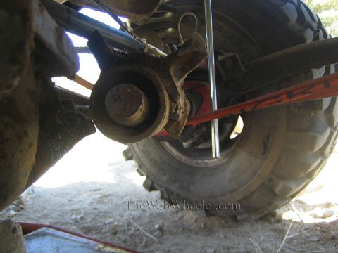 1990 Ford Bronco - Close underbody view of a 4x4's front axle knuckle and exposed stub shaft, orange recovery strap and oversized tire behind.