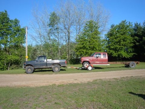 1985 Ford F-150 - Two pickup trucks are parked side-by-side on a dirt road with trees and a utility pole in the background.