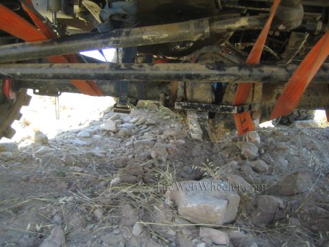 1990 Ford Bronco - Underbody of a 4x4 showing the axle tube and frame rails braced with orange straps, over a bed of rocks and dry straw.