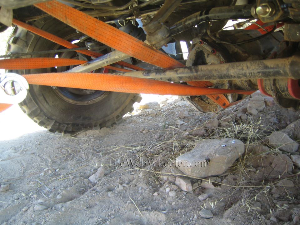 Underside of a 4x4 with orange recovery straps crossing tire and axle, rock below