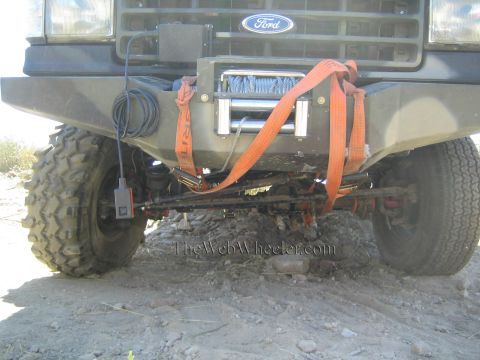 1990 Ford Bronco - Front bumper of a Ford 4x4 with a cable winch and orange recovery straps strung across the exposed axle on rocky ground.