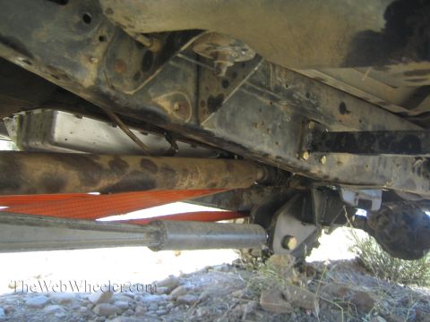 1990 Ford Bronco - Underbody view of a vehicle showing the frame rails, fuel tank, driveshaft and an orange strap over rocky ground.