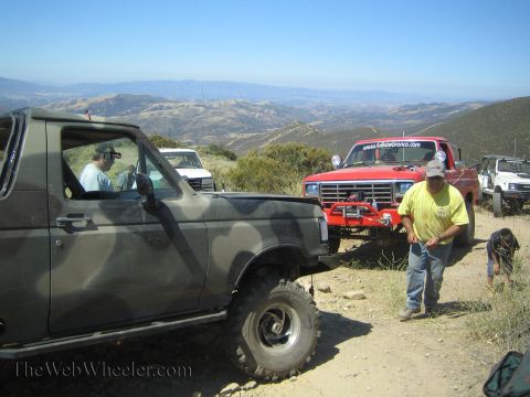 1990 Ford Bronco - camouflage Ford Bronco and a red Ford truck park on a dirt mountain ridge as a man in a yellow shirt walks alongside.