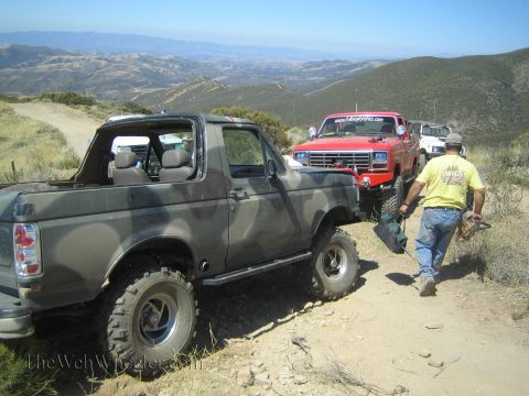 1990 Ford Bronco - topless camouflage Ford Bronco sits on a dirt ridge trail beside a red Ford truck, a man in yellow carrying gear nearby.