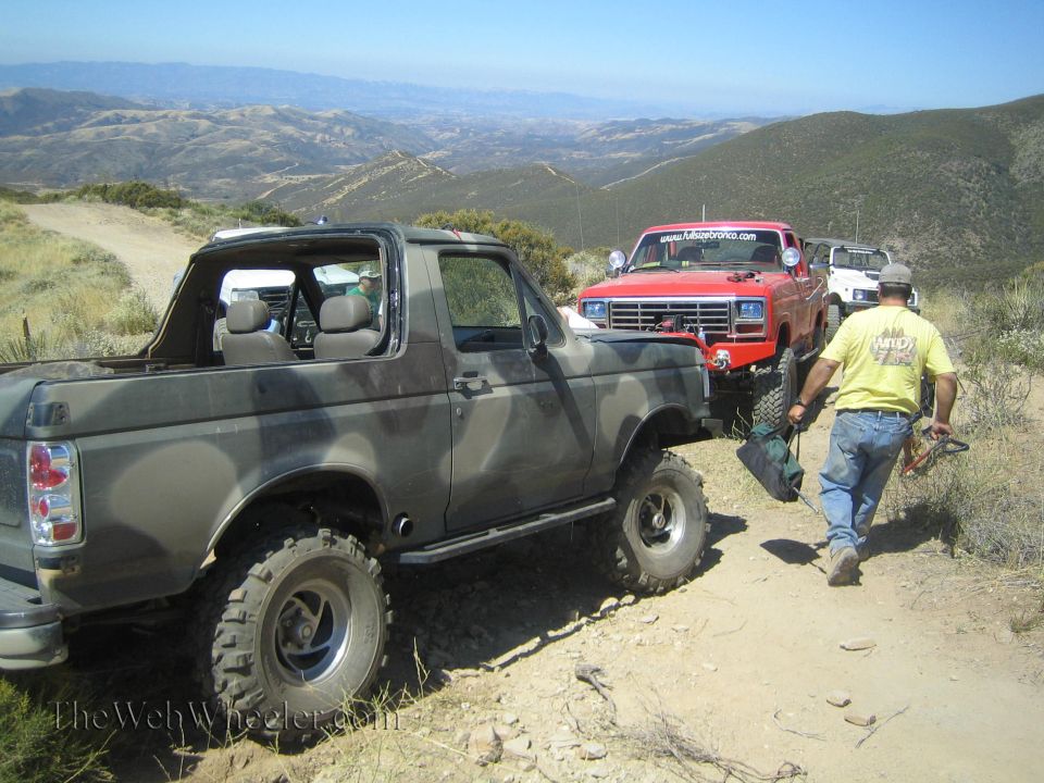 Camouflage Ford Bronco with top off beside a red Ford truck on a dirt ridge