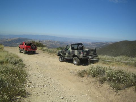 1990 Ford Bronco - camo-painted topless 4x4 and a red Jeep travel a dusty ridgeline dirt trail above rolling green hills.