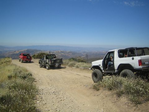 1990 Ford Bronco - white open-top 4x4 with a driver inside sits on a hilltop dirt trail beside a camo truck and distant red Jeep.