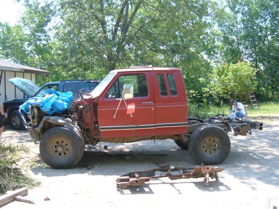 Red extended-cab Ford pickup stripped to cab and frame in a gravel driveway, bed and front clip removed