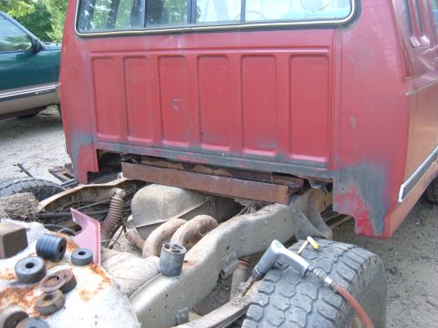 1985 Ford F-150 - The rear section of a red truck cab and chassis is shown, with visible rust and suspension parts. Another vehicle is partially visible to t…