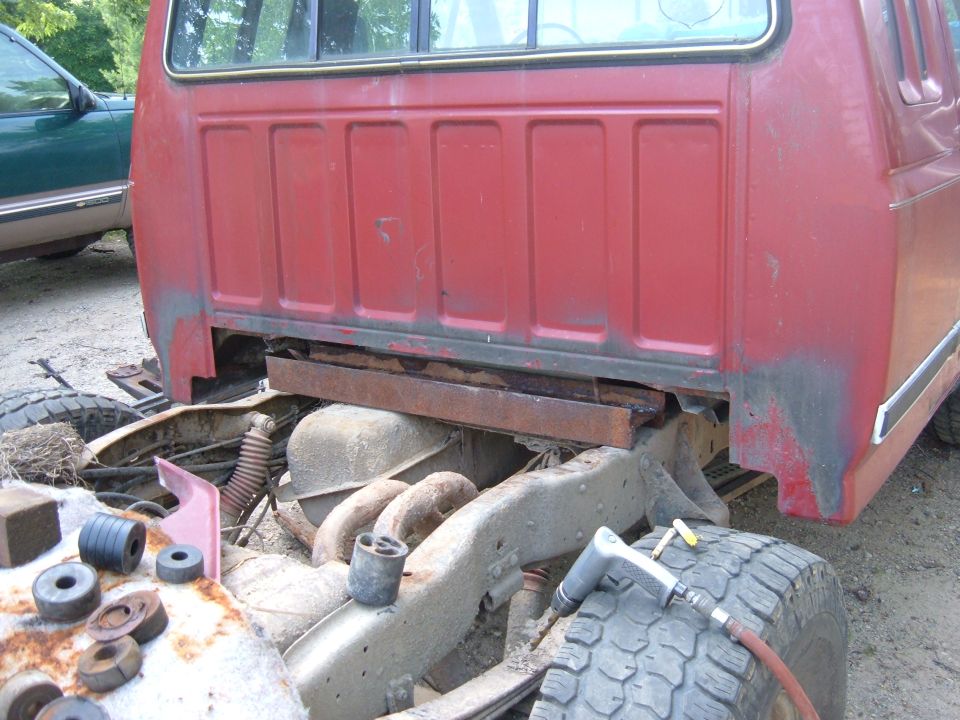 Red truck cab and chassis with visible rust and suspension components. A green vehicle is partially visible on the left