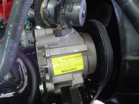 1984 Ford Bronco - Close-up of a remanufactured power steering pump newly installed on an engine, its bare aluminum body wearing an upside-down yellow CAUTION…
