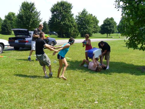 1991 Ford Probe - A group of people are playing with water guns in a park next to a blue sedan with its trunk open.
