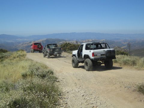 1990 Ford Bronco - lifted white open-top 4x4 climbs a dirt ridgeline trail, linked by a strap toward a red Jeep and camo truck ahead.