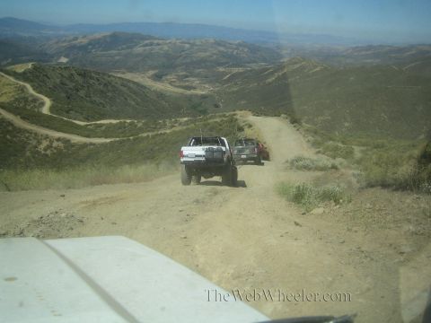 1990 Ford Bronco - Seen over a vehicle hood, a white truck and another vehicle descend a steep winding dirt road into a hilly valley.