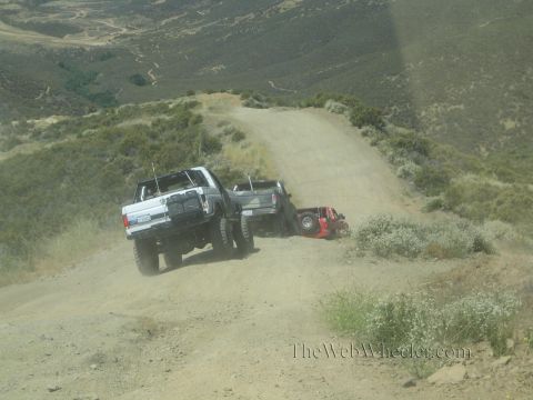 1990 Ford Bronco - lifted white truck connected by a recovery strap works to pull a vehicle off the brushy edge of a hillside dirt road.