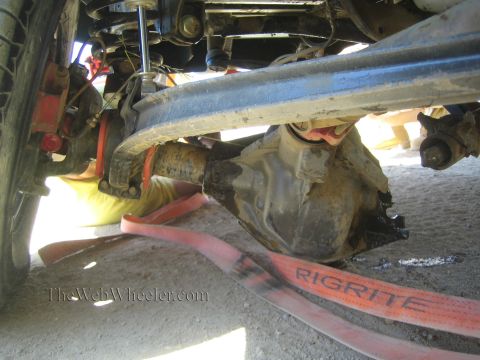1990 Ford Bronco - Low-angle underbody shot showing a front axle, leaf spring, driveshaft and an orange RIGRITE strap on sandy ground.