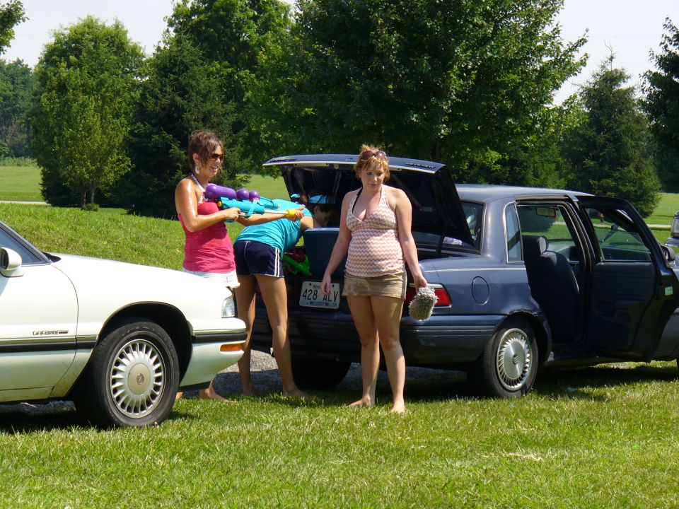 Two women with water guns stand by a blue sedan with an open trunk, next to a white car