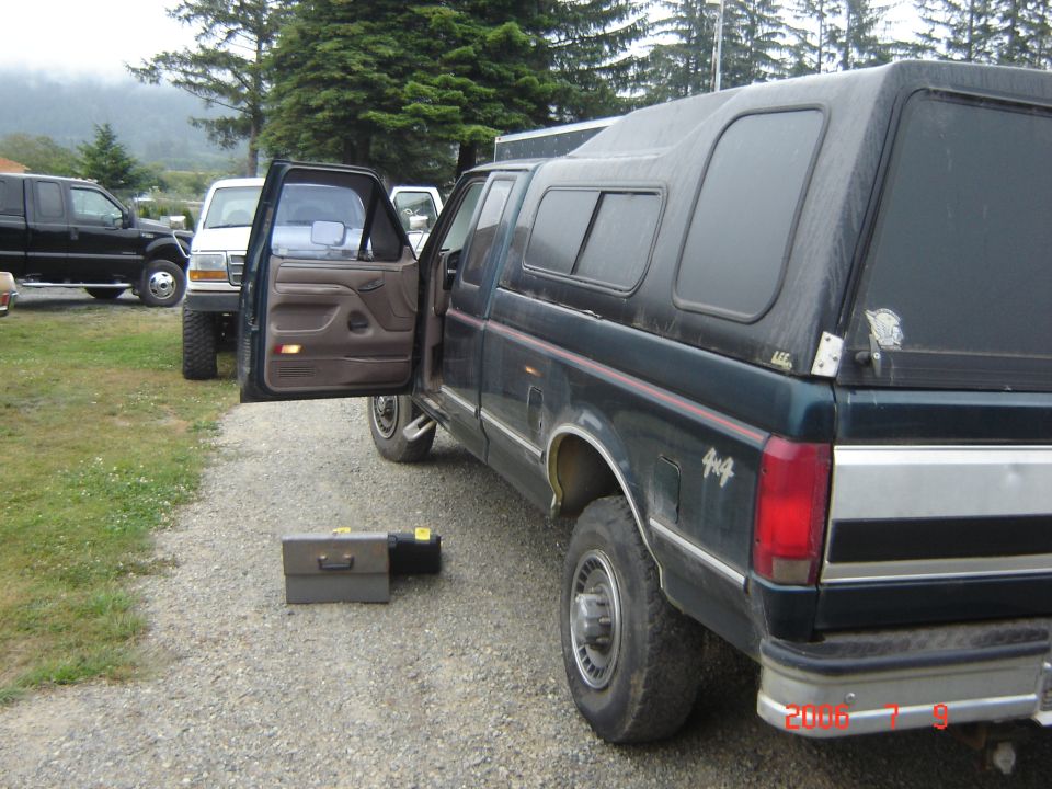 Dark green pickup truck, 4x4 badge on fender, camper shell, open driver door, toolbox on ground