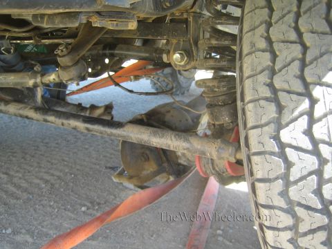 1990 Ford Bronco - Underbody view of a lifted off-road vehicle's front axle, steering linkage and a large all-terrain tire with an orange strap.