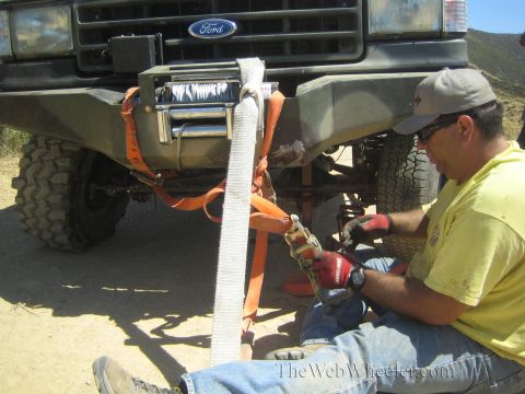 1990 Ford Bronco - seated man in a yellow shirt rigs an orange strap to the winch on a Ford 4x4's front bumper on a dirt trail.