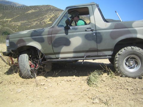 1990 Ford Bronco - Side profile of a camo-painted open-top 4x4 with a driver inside, tilted on rocky dirt with a hillside behind.