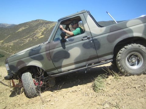 1990 Ford Bronco - camo-painted topless 4x4 with a smiling driver inside sits tilted on rocky ground below brown rolling hills.