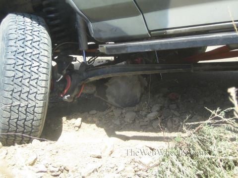 1990 Ford Bronco - Low underbody shot of a vehicle's solid axle, leaf spring and large tire resting over loose rock and dirt.
