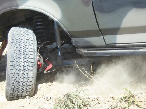 1990 Ford Bronco - Close-up of a lifted vehicle's front wheel, blue shock and rock-slider step throwing dust as it works through dirt.