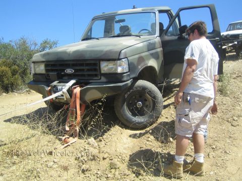 1990 Ford Bronco - man in a white shirt stands beside a camo-painted Ford 4x4 as its front winch strap runs taut across the dirt trail.