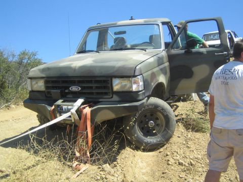 1990 Ford Bronco - camo-painted Ford 4x4 sits with doors open and front winch line out while people in T-shirts stand beside it on the trail.