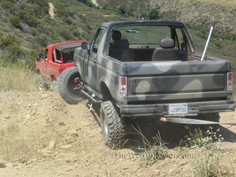 1990 Ford Bronco - Rear view of a lifted camo open-top truck showing a California plate, parked on a hillside trail next to a red truck.